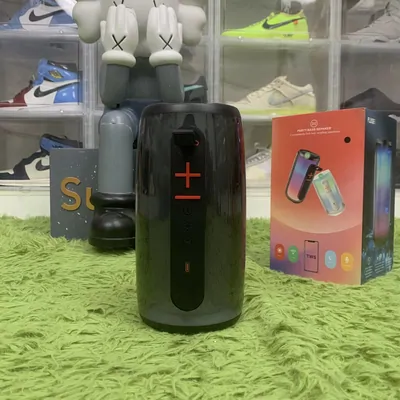 A black cylindrical bluetooth speaker standing on green turf, with sneakers and toy figures on the shelves behind it.
