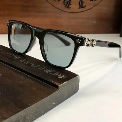 Black square-framed glasses with a metal cross detail on the temple, photographed on a wooden tray.