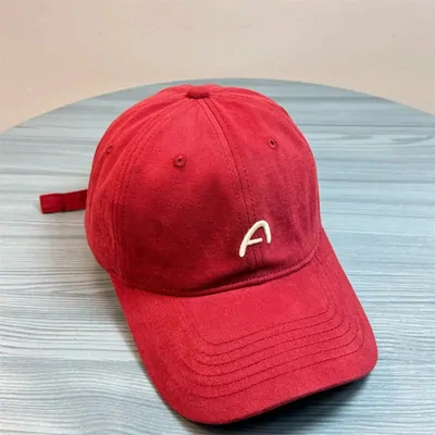 A red suede-look baseball cap with a white letter A embroidered on the front, resting on a pale blue surface.