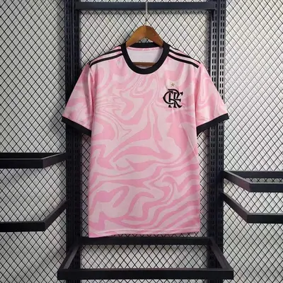 A pink football shirt with a white marbled swirl pattern and a club crest, on a wooden hanger against a dark mesh wall.