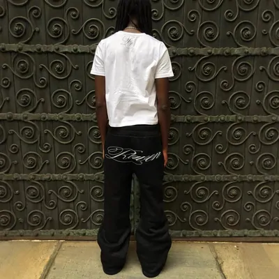 A person seen from behind against an ornate iron gate, in a white tee and black wide-leg sweatpants with a script logo down the leg.